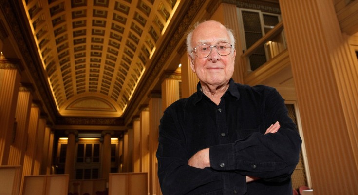 Peter Higgs stands with his arms crossed in a large hall