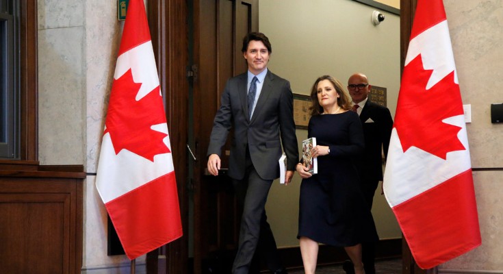 A photo of Canadian prime minister Justin Trudeau and finance minister Chrystia Freeland walking through a doorway flanked by large Canadian flags. A man in a suit and glasses walks behind them