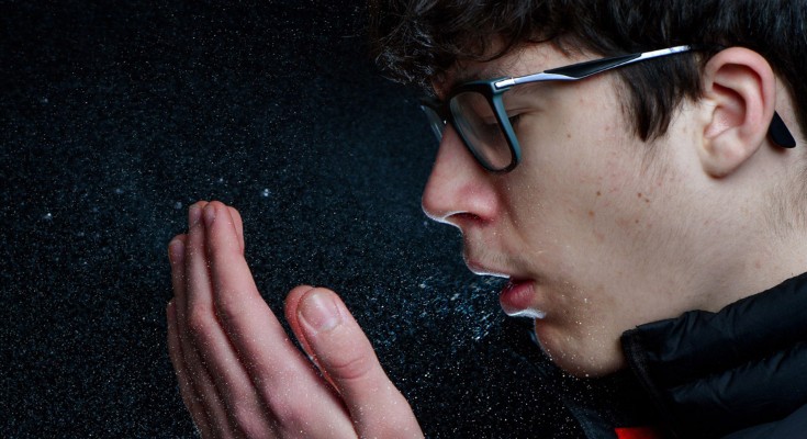 A high speed photograph illustrates the many droplets expelled into the air by a sneezing teenage boy