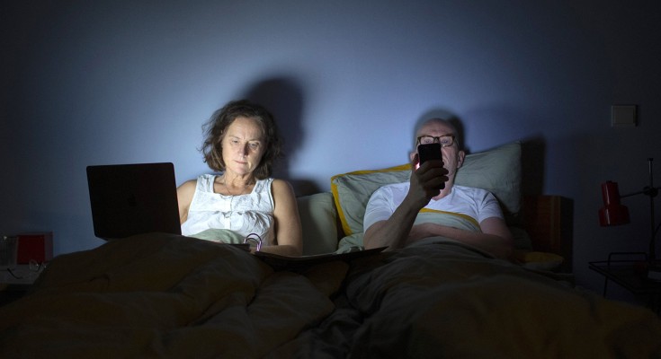 A man and woman sit up in bed in a dark room with a blue wall and green bed linen. She has a laptop on her lap throwing light up on her face. He holds a mobile phone in front of his face. There are both in their 60-70s and wearing white pyjamas.