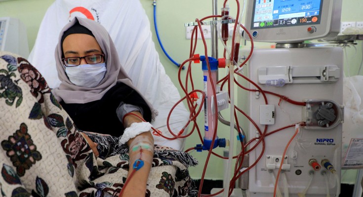 A person sits up in a hospital chair. They are wearing a headscarf and face mask, and are covered in a floral patterned blanket. They have a bandage on their hand, and in their forearm they have a cannula, connected to a dialysis machine