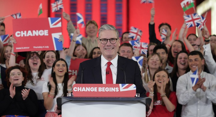 New Prime Minister Sir Keir Starmer stands in front of a lectern with a sign reading "change begins" with a Union flag. Behind him, crowds cheer