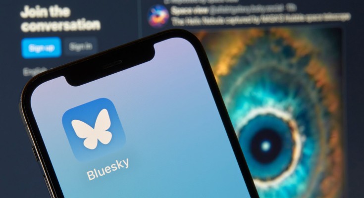 A smartphone displaying the icon for the Bluesky social media app, in front of a computer screen featuring a Bluesky feed