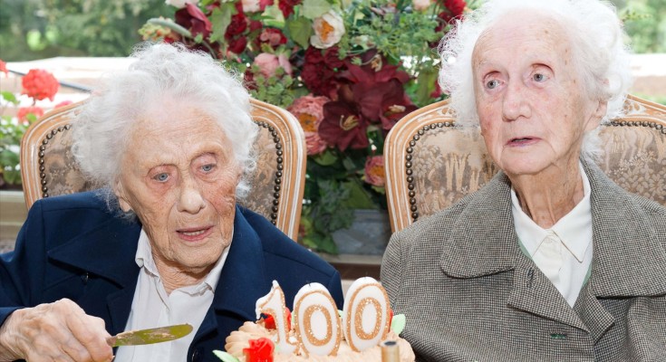 Two elderly women sit in chairs next to each other, in front of them there is a cake with candles reading 100
