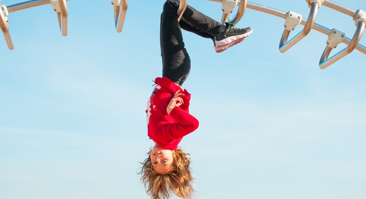 A child hanging upside down by her knees on a climbing frame looking at the camer
