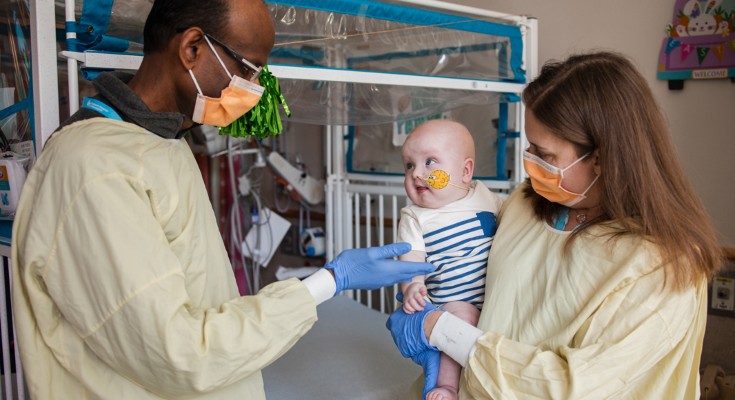 A woman holds a baby, who has a breathing tube. A man holds his hand out to the baby