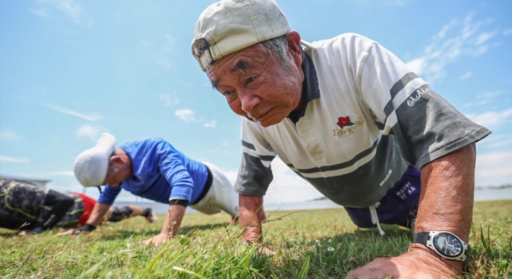 An elderly man is in the plank position on some grass, with others in the same position in the background