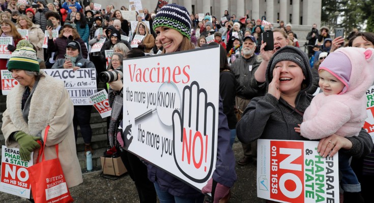A group of protesters hold signs, the foremost sign says "Vaccines, the more you KNOW, the more you NO!"