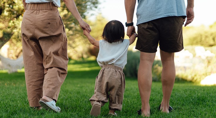 A small child walks away from the camera, an adult holding their hand on each side