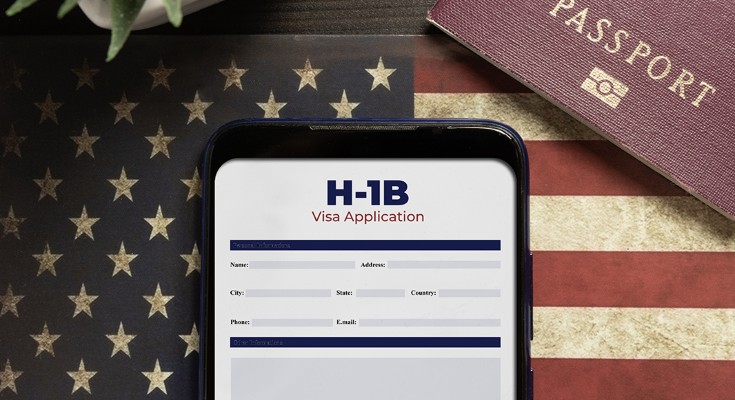 A phone with a H-1B application on the screen sits atop an American flag. There is a passport in the corner
