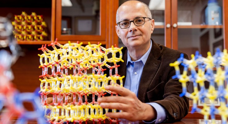 A portrait of Omar Yaghi sitting at a wooden desk with a red and yellow model