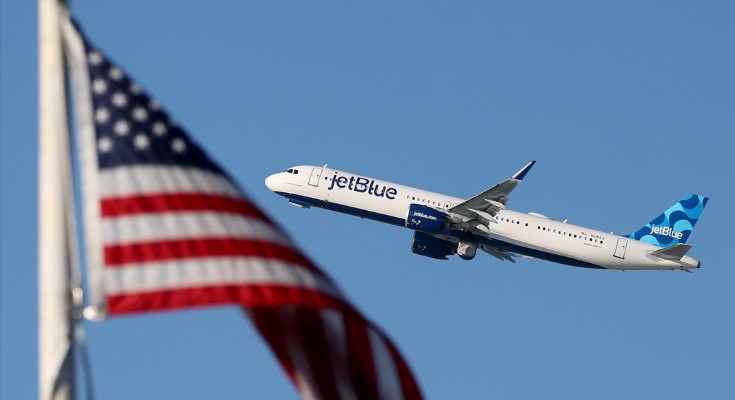 A Jet Blue plane flies upwards, with an out of focus USA flag in the foreground