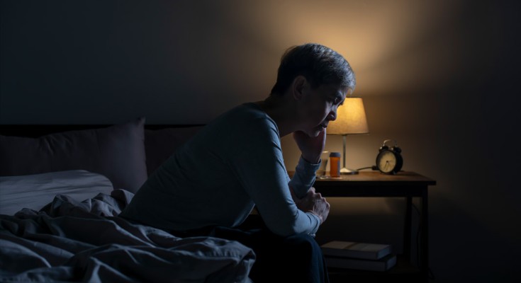 A person sits on the end of a bed, head in hand. It is dark and there is a bedside lamp on