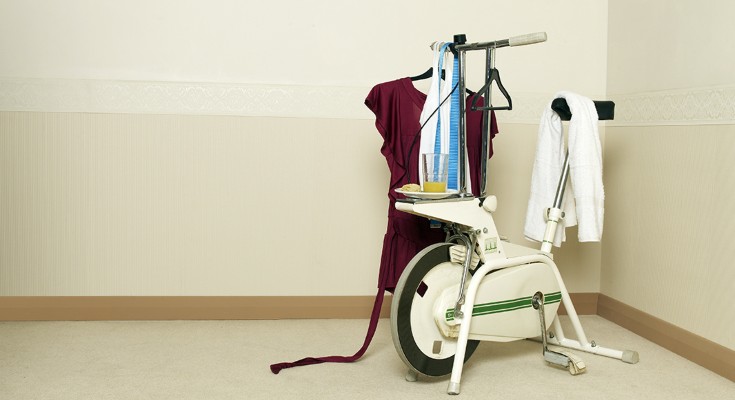 An exercise machine sits in the corner of a room with clothes hanging off it, and a plate with half-eaten food, and a nearly empty glass of juice
