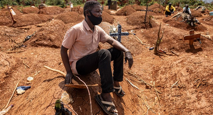A man wearing a face covering sits by a newly dug grave in a cemetery in the DRC