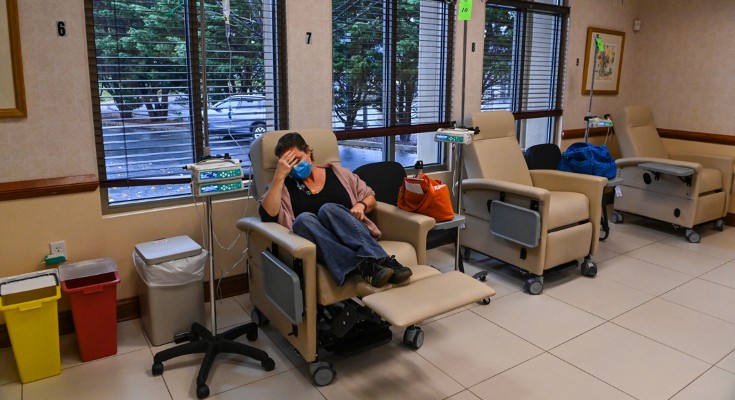 A woman is sat in a recliner chair, receiving chemotherapy. She is wearing a face covering and has her head in her hand