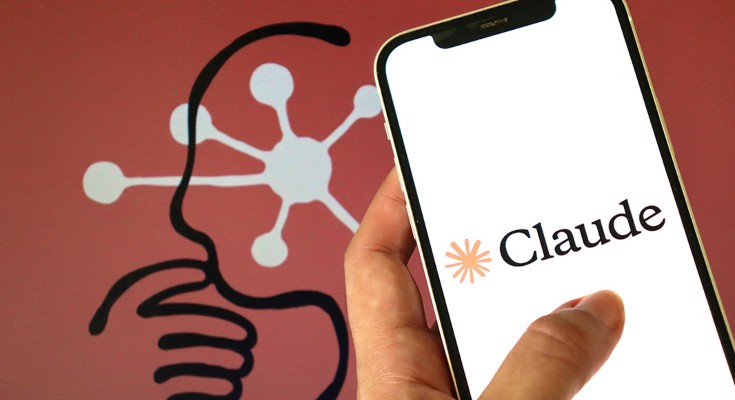 A close-up view of a hand holding a smartphone, with the Claude logo on the screen, and a graphic of an amorphic thinking head in the background