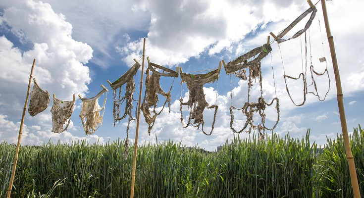 Various pieces of handmade netting or woven fabric hanging on a wooden frame outdoors against a cloudy sky and tall green grass in the background.