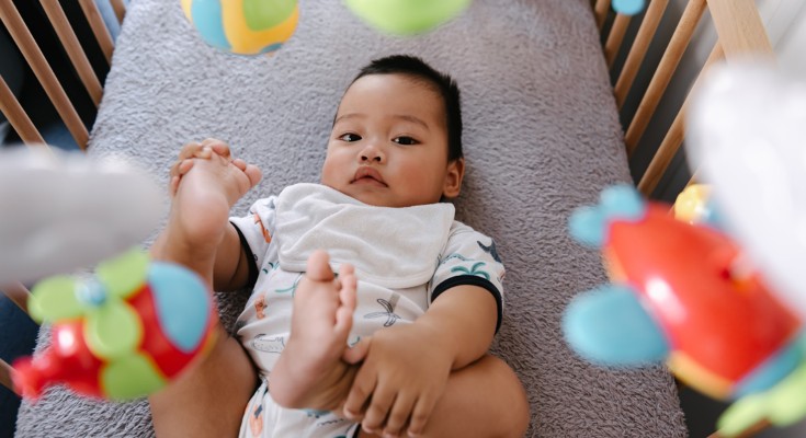 A baby looks up at the camera from it's crib, with a mobile visible above them. They play with their feet