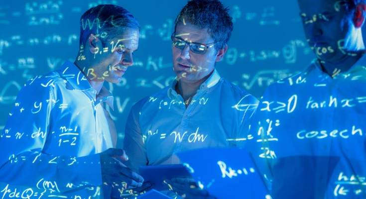 Three men stand looking at sheets of paper while equations are projected on them 