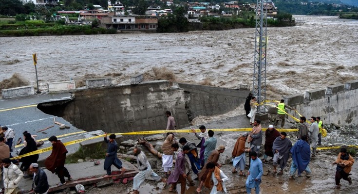 A mass of people walk to and fro in front of a paved road that has been ripped apart by flash floods. A river, overflowing its banks, rushes along and over the road.