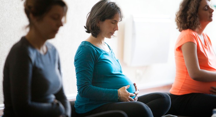 Three pregnant women sit cross-legged on yoga mats while holding their bumps