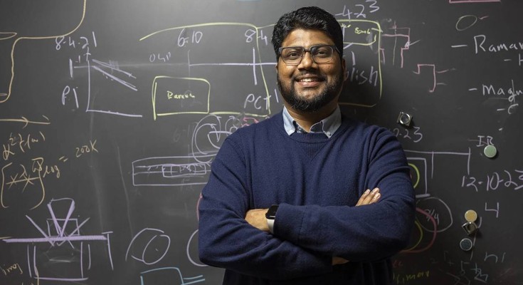 Physicist Ranga Dias, wearing a collared shirt underneath a sweater, stands in front of a blackboard with notations on it. He wears glasses and has his arms crossed in front of him as he smiles for the camera.