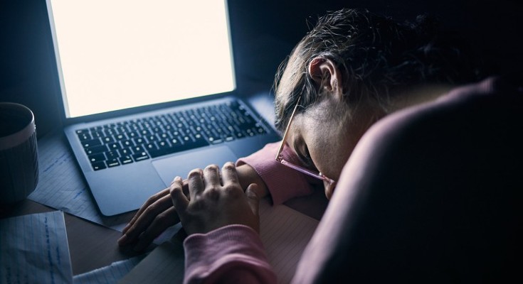 A woman sleeps at a desk, slumped on an illuminated laptop