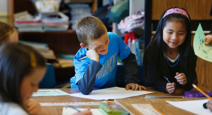 A primary school age boy and girl smile as they sit at a desk in a classroom while working on maths problems
