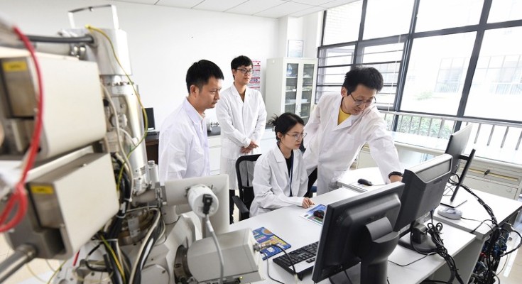 Three young Chinese scientists in lab coats, looking at a computer monitor, one of them is seated and there is a fourth older scientists offering advice.