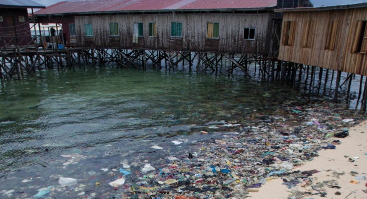 Multicoloured plastic pollution collects at the shoreline of a beach with a wooden building on stilts over the water in the background.